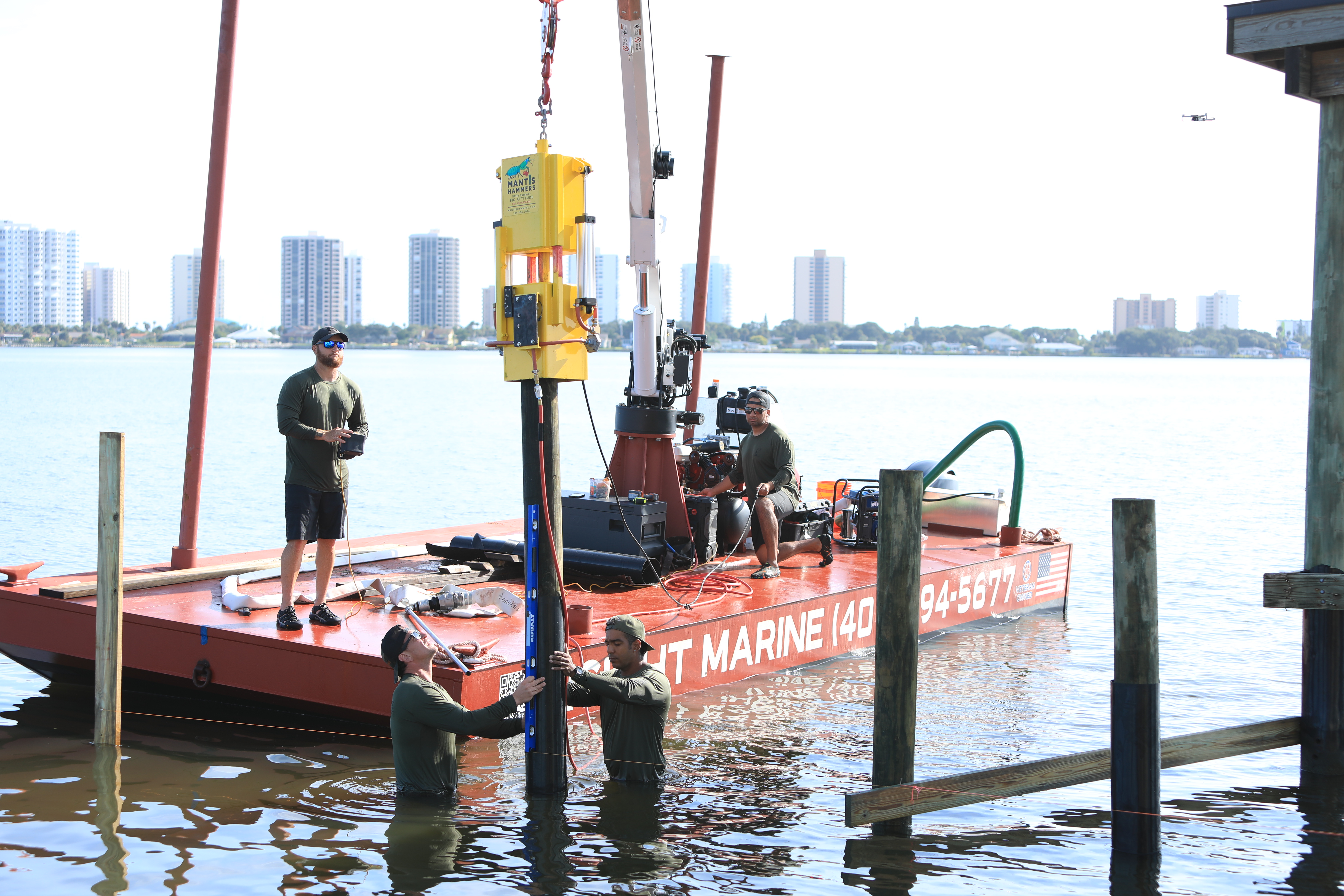Crew driving a timber pile with a hydraulic hammer hung from the barge crane